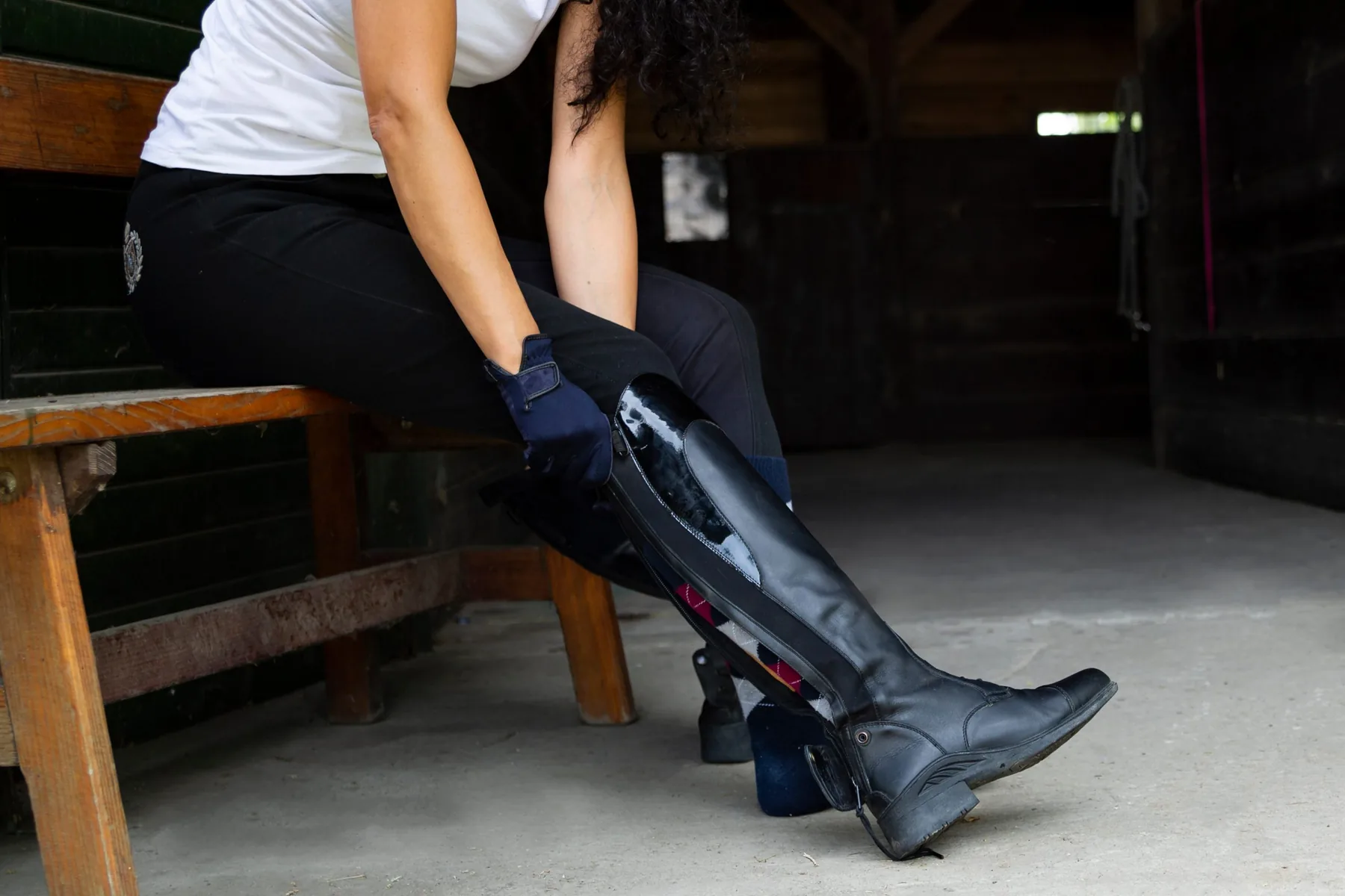 Equestrian rider putting on tall black riding boots in a tack-room setting